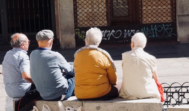Zdjęcie czterech starszych osób siedzących na murku tyłem. Po lewej stronie znajduje się dwóch panów, po prawej dwie panie.