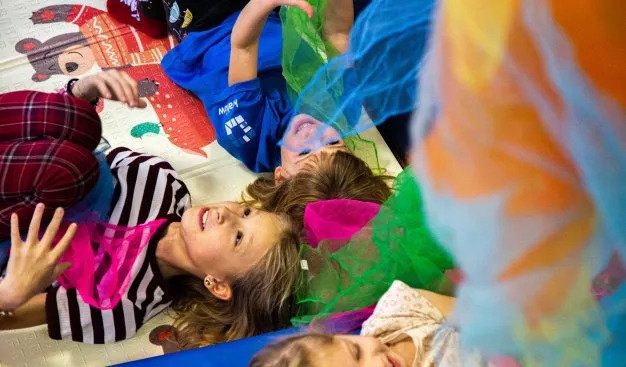 Dzieci leżą na matach zbliżone do siebie głowami. Podrzucają kolorowe  chusty z cienkiego materiału.