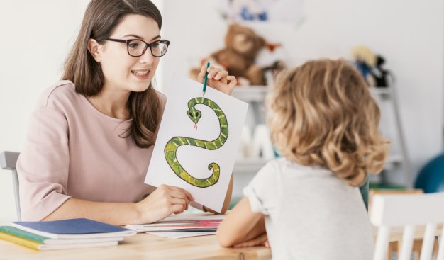 Kobieta siedzi na przeciwko dziecka przy stole. Pokazuje mu kartkę z narysowanym wężem, układającym się w literę "S". Ćwiczą dykcję.