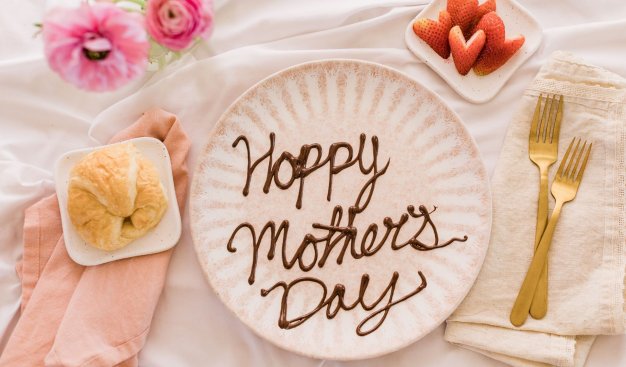 Zdjęcie przedstawiające talerz z napisem: Happy Mother's Day pośrodku, w otoczeniu sztućców, rogalika oraz truskawek wyciętych w serca