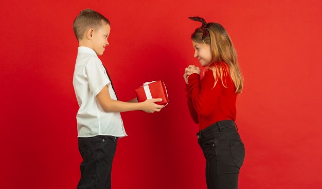 Zdjęcie dwójki dzieci stojących na czerwonym tle, chłopiec trzyma pudełko prezentowe, które wręcza dziewczynce