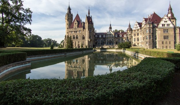 Zdjęcie przedstawia gmach zamku w Mosznej, odbijający się w mieszczącej się przed nim fontannie.