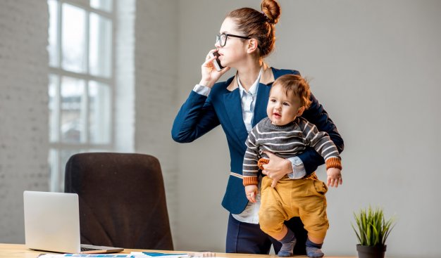 Kobieta ubrana w garsonkę, stojąca przed biurkiem, na którym leży otwarty laptop. W jednej ręce trzyma małe dziecko, w drugiej telefon komórkowy, przez który rozmawia.
