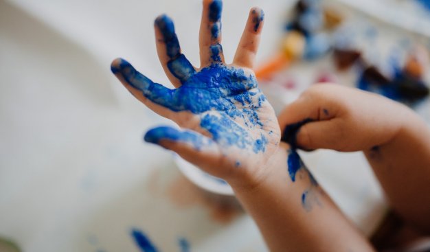 Na pierwszym planie znajduje się dłoń kilkuletniego dziecka. Na ręce dziecka odbita jest niebieska farba. Rozmazane tło zdjęcia przedstawia farby oraz białą kartkę lub płótno.