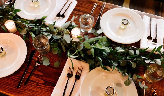 Zdjęcie stołu nakrytego białą zastawą, z zieloną dekoracją leżącą po środku stołu.