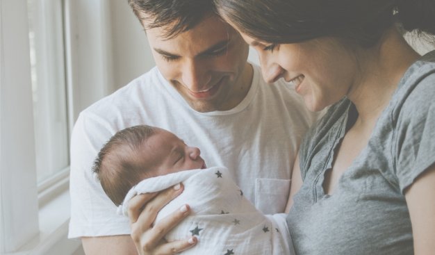 Kobieta i mężczyzna z malutkim dzieckiem. Kobieta trzyma niemowlę owinięte w jasną pieluszkę. rodzice z uśmiechem spoglądają na dziecko.