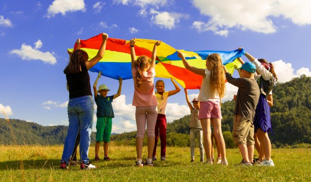 Zdjęcie przedstawia grupę ludzi wznoszących rozłożone, okrągłe i kolorowe płótno służące do zabawy. W tle słoneczna pogoda.