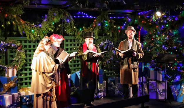 Zdjęcie przedstawiające grupę osób śpiewających, trzymają oni śpiewniki i są ubrani w świąteczne stroje, na drugim planie znajdują się bożonarodzeniowe dekoracje w tym choinki z bąbkami.