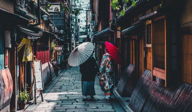 uliczka w azjatyckim mieście, idące po niej dwie postaci odwrócone tyłem, obie z parasolami
