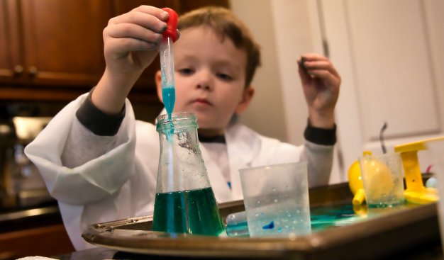 Zdjęcie przedstawiające małego chłopca w białym fartuchu wykonującego doświadczenie chemiczne przy użyciu pipety i zlewki.