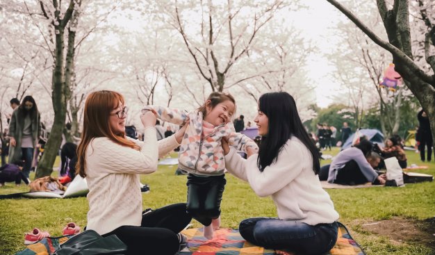 Na zdjęciu widać dwie azjatyckie kobiety bawiące się z azjatycką dziewczynką na pikniku. Osoby siedzą na kocu w parku w otoczeniu innych piknikowiczów.