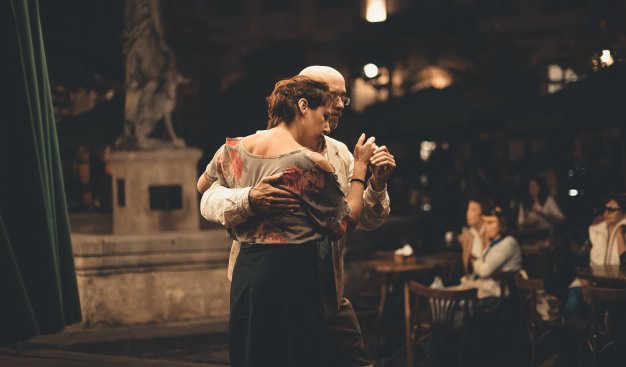 Na zdjęciu kobieta tańcząca w objęciu mężczyzny wieczorową porą, oświetleni przez lampy. Na drugim planie widać parę siedząca przy stoliku oraz pomnik.