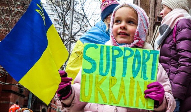 Mała dziewczynka z flagą ukrainy w jednej dłoni a w drugiej z kartką "Support Ukraine"