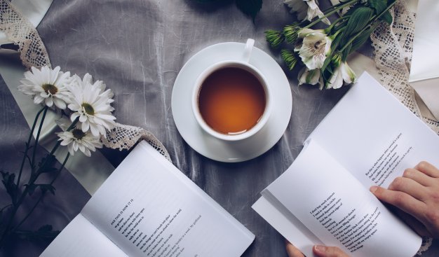Zdjęcie przedstawiające dwie otwarte książki i filiżankę z herbatą w otoczeniu kwiatów