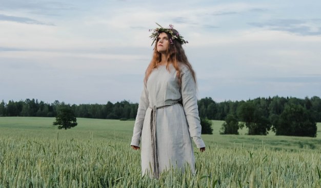 Młoda kobieta w prostym, ludowym stroju i wianku na głowie, stoląca na zielonym polu.