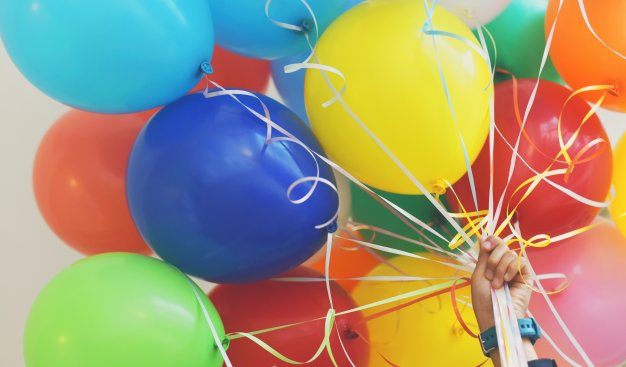 Zdjęcie kolorowych balonów z helem, trzymanych za kolorowe wstążeczki przez jedną, dziecięcą dłoń