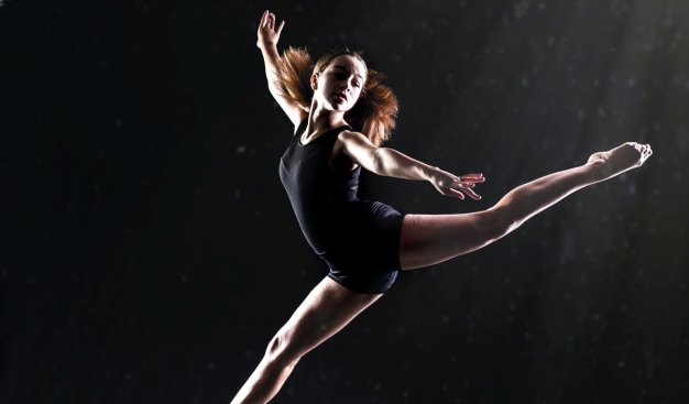 Dziewczyna w obcisłym body, w tanecznym wyskoku z szeroko rozłożonymi nogami, na ciemnym tle, oświetlona od góry jasnym, punktuwym światłem