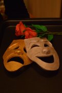 Na czarnej podkładce leżą róże dwie maski teatralne. Przed różami leżą maski opierające się o siebie. Jedna z nich jest pozłacana i jest smutna, a druga szczęśliwa i jest biała.