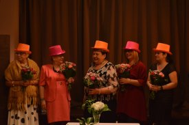 Wnętrze Klubu Dukat. Na zdjęciu przed sceną stoją członkinie Klubowej Grupy Poetica.  Wszystkie trzymają małe bukiety kwiatów. Są ubrane w sukienki oraz w kolorowe kapelusze