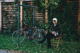 Fotografia przedstawiająca starszego Pana, który siedzi w ogrodzie, na krześle. Obok niego stoi jest rower, oparty o ogrodową suszarkę na pranie.