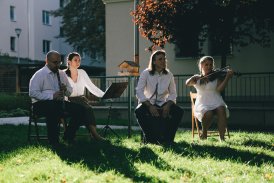 Scena z plenerowego spektaklu, przedstawiająca czteroosobową grupę artystów, którzy grają na instrumentach. Siedzą oni w ogrodzie, na krzesłach. W tle są budynki mieszkalne.