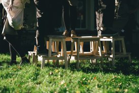 Scena z plenerowego spektaklu, przedstawiająca części nóg ludzi, stojących na krzesłach, w ogrodzie.