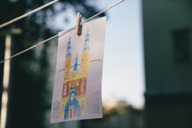 Fotografia przedstawiająca rysunek dziecka, wiszący na lince od suszarki na pranie. Przyczepiony jest spinaczem. Rysunek przedstawia Kościół z Matką Boską. W tle jest natura.