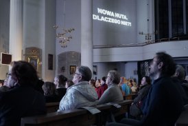 Zdjęcie przedstawiające publiczność koncertu, siedzącą w ławach kościelnych. W tle znajduje się napis Nowa Huta. Dlaczego nie?!