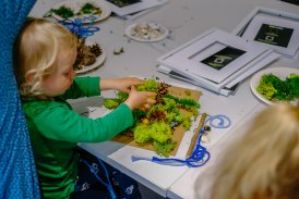 Zdjęcie z warsztatów tworzenia obrazów z mchu, przedstawiające kilkuletnie dziecko siedzące przy stole i przyklejające zielony mech na kartonowy podkład.
