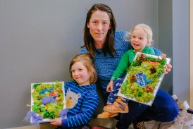Zdjęcie przedstawia mamę z dziećmi wraz z ich pracami. Dzieci wtulone w matczyne ramiona przedstawiają wykonane prace z mchu.