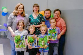 Na zdjęciu grupa uśmiechniętych ludzi- czterech dorosłych i czworo dzieci. Dzieci prezentują wykonane przez siebie obrazy z mchu.