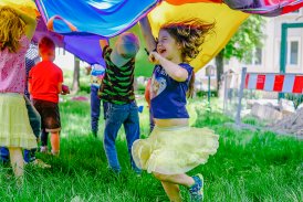 Na zdjęciu widać dzieci bawiące się pod kolorowym płótnem. Na głównym planie rozbawiona dziewczynka w żółtej spódniczce .