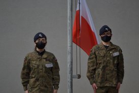 Dwoje młodych ludzi w mundurach wojskowych stoją na baczność pod masztem z biało-czerwoną flaga narodową.