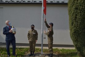 Dwoje młodych ludzi w mundurach wojskowych - jedna z nich wciąga flagę na maszt, druga stoi stoją na baczność pod masztem.
