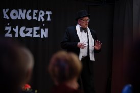 Fotografia przedstawiająca starszego mężczyznę, stojącego na scenie, w Klubie Dukat. Ubrany jest w garnitur. W tle biały napis " Koncert Życzeń", przypięty na czarnym tle.