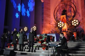 Wnętrze kościoła. Przed ołtarzem oświetlonym ciepłym, pomarańczowym światłem stoi trzech mężczyzn. Mężczyźni trzymają mikrofony przy ustach i śpiewają. Z boku, po prawej stronie, siedzi inny mężczyzna i gra na pianinie cyfrowym.