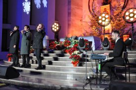 Wnętrze kościoła. Przed ołtarzem oświetlonym ciepłym, pomarańczowym światłem stoi trzech mężczyzn. Mężczyźni trzymają mikrofony przy ustach i śpiewają. Z boku, po prawej stronie, siedzi inny mężczyzna i gra na pianinie cyfrowym.