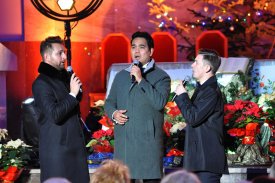 Na pierwszym planie stoi trzech mężczyzn. Mężczyźni śpiewają trzymając mikrofony przy ustach. Za nimi widać dekoracje bożonarodzeniowe oraz część ołtarza.