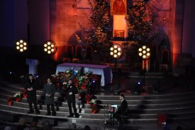 Wnętrze ciemnego kościoła. Przed udekorowanym bożonarodzeniowo ołtarzem stoi trzech mężczyzn. Jeden z nich trzyma przy ustach mikrofon i wygląda jakby śpiewał. Obok siedzi inny mężczyzna i gra na pianinie cyfrowym.