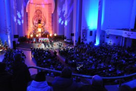Wnętrze ciemnego kościoła oświetlonego niebieskim światłem. W ławkach siedzi mnóstwo ludzi słuchających koncertu. Przed ołtarzem oświetlonym ciepłym, pomarańczowym światłem stoi trzech śpiewających mężczyzn. Obok nich siedzi czwarty i gra na pianinie cyfr