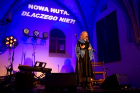 Ciemne, gotyckie pomieszczenie oświetlone fioletowo-niebieskim światłem. Na scenie stoi kobieta i przemawia przez mikrofon. Dookoła niej stoją słabo świecące reflektory, pianino cyfrowe i krzesło. Za nią na ścianie wyświetlony jest napis "Nowa Huta. Dlacz