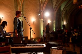 Wnętrze ciemnej gotyckiej sali. Salę słabo oświetlają kinkiety zawieszone wzdłuż jednej ściany. Na scenie stoi kobieta i śpiewa przez stojący przed nią mikrofon. Obok siedzi mężczyzna i gra na gitarze. Przed sceną siedzi pogrążona w mroku widownia.