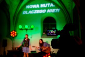 Ciemna gotycka sala. Na pierwszym planie widać kamerę z podglądem kamerującą scenę. Na scenie w tle stoi kobieta i śpiewa przez mikrofon. Obok siedzi mężczyzna i gra na gitarze. Scena oświetlona jest zielonym światłem. Za nią na ścianie wyświetlony jest n