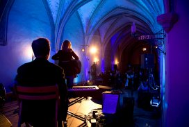 Ciemna gotycka sala oświetlona niebieskim światłem. Na scenie, przodem do widowni, stoi kobieta i gra na gitarze. Za nią siedzi na krześle mężczyzna. Przed nią widać zacienioną widownię.