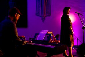 Ciemna sala, słabo oświetlona fioletowym światłem. Na scenie stoi kobieta. Jest lekko odwrócona od mikrofonu stojącego przed nią. Za nią siedzi mężczyzna i gra na pianinie cyfrowym.
