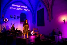 Ciemna, gotycka sala oświetlona fioletowym światłem. Na scenie stoi kobieta. Przed nią znajduje się mikrofon na stojaku. Za kobieta siedzi mężczyzna i gra na pianinie cyfrowym. Na ścianie za sceną jest wyświetlony napis "Nowa Huta. Dlaczego nie?".