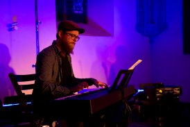 Ciemna sala oświetlona słabym, fioletowym światłem. Na scenie siedzi mężczyzna w berecie i gra na pianinie cyfrowym.