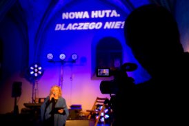 Ciemna sala oświetlona niebieskim światłem. Na scenie stoi kobieta oświetlona bardzo jasnym, ciepłym światłem. Kobieta przemawia przez mikrofon. Na pierwszym planie widać człowieka z kamerą z podglądem kamerującego scenę.