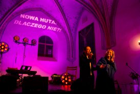 Ciemna gotycka sala, oświetlona różowym światłem. Dwie kobiety stoją na scenie. Obie trzymają w rękach mikrofony. Jedna z nich przemawia, zwrócona twarzą do widowni. Druga spogląda na nią. Obie kobiety się uśmiechają. Za nimi na ścianie napis "Nowa Huta. 