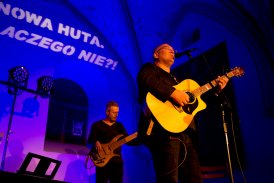 Ciemna, gotycka sala oświetlona granatowym światłem. Na scenie stoi dwóch mężczyzn grających na gitarach. Mężczyzna z przodu śpiewa do stojącego przed nim mikrofonu.